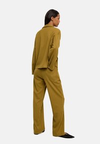 Musztardowy garnitur w paski o luźnym kroju. Zawiera kurtkę bez kołnierzyka i spodnie o szerokich nogawkach. Całość dopełniają czarne, spiczaste baleriny.