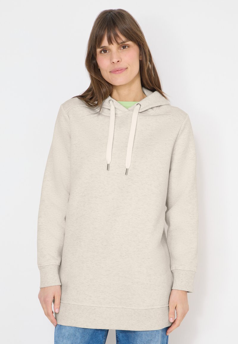 Femme aux cheveux bruns portant un sweat à capuche beige clair avec cordons et un jean bleu, debout devant un fond blanc.