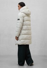 Cappotto imbottito di colore grigio chiaro, con cappuccio, design trapuntato e etichetta di stoffa nera sulla manica sinistra. Abbinato a pantaloni larghi di colore scuro.