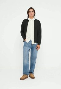 Jauns vīrietis melnā jaku, baltu kreklu, zilas džinsa bikses un bēšas kurpes, stāvot pret baltu fonu.