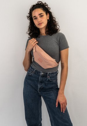 Fanny pack in soft pink fabric with a smooth texture and zip closure, worn over a gray t-shirt and high-waisted blue jeans.