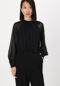 Zwarte blouse met doorzichtige lange mouwen, hoge halslijn en een gestrikt detail bij de kraag. Gecombineerd met hooggetailleerde zwarte broek.
