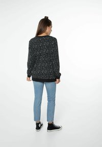 Maglione nero con macchie bianche, scollatura rotonda e maniche con polsini. Indossato con jeans azzurri e sneakers alte nere.