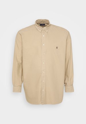 Chemise beige à manches longues, en tissu de coton doux. Elle présente un col pointu et un petit logo brodé sur la poitrine gauche.