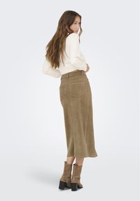 Falda de ante verde oliva con longitud hasta la mitad de la pantorrilla, que presenta bolsillos y detalles de costura. Combinada con botas cortas de tacón marrón texturizadas.