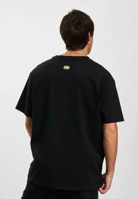 Black cotton t-shirt with a relaxed fit, short sleeves, and a small yellow logo centered on the back neck area. Smooth texture.