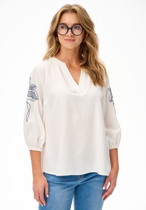 Kobieta o długich blond włosach i okrągłych okularach, ubrana w białą bluzkę z niebieskim kwiatowym haftem na rękawach oraz niebieskie dżinsy.