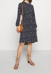 Granatowa sukienka w kwiatowy wzór, z zapięciem na guziki, długimi rękawami i ściągniętą w talii. Połączona z brązowymi sandałami na paski i okrągłą torbą.