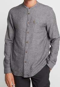 Hombre vestido con una camisa de botón gris de manga larga con cuello mandarín y bolsillo en el pecho, combinado con pantalones oscuros, con las manos relajadas a los lados.