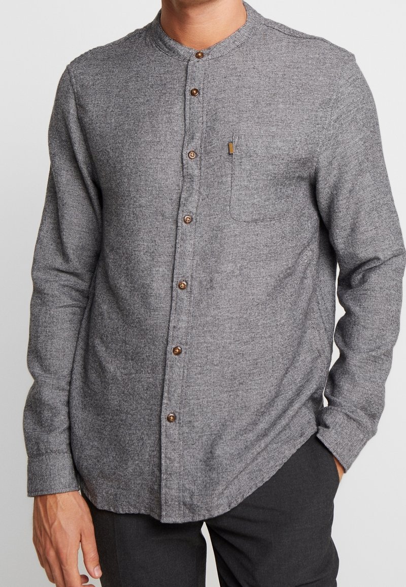 Hombre vestido con una camisa de botón gris de manga larga con cuello mandarín y bolsillo en el pecho, combinado con pantalones oscuros, con las manos relajadas a los lados.