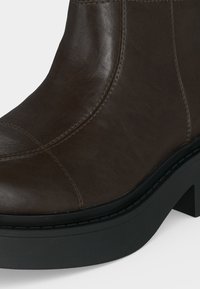 Gros plan d'une botte cheville en cuir marron avec des coutures visibles et une semelle noire épaisse avec talon bloc.