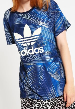 Person iført en blå Adidas-trøje med hvide konturlinjemønstre og et nederdel med leopardprint.