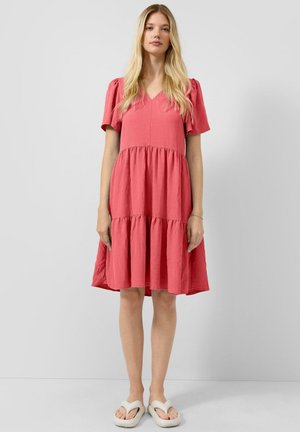 Jeune femme aux longs cheveux blonds portant une robe corail rose à étages avec des manches bouffantes et des sandales blanches à plateforme, debout devant un fond uni.