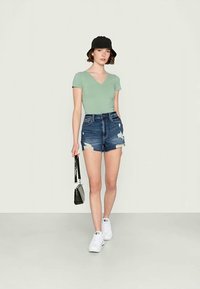 T-shirt verde em decote em V, calções de ganga desgastados, sapatilhas brancas e um chapéu de balde preto, segurando uma pequena bolsa preta.