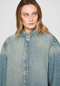Camicia in denim blu chiaro con chiusura a bottoni, colletto classico e vestibilità over-size. Presenta un tessuto testurizzato e una leggera scoloritura.