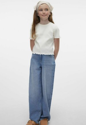 Hvid ribbet kortærmet top med bølget kant, parret med brede blå denim jeans. Modellen bærer et blomsterhovedklæde og brune sandaler.