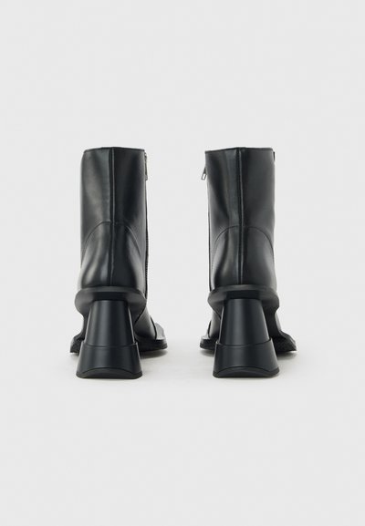 Μαύρα δερμάτινα μποτάκια αστραγάλου με χοντρές διαμορφωμένες τετράγωνες φτέρνες και πλευρικά φερμουάρ, εμφανίζονται από πίσω σε λευκό φόντο.