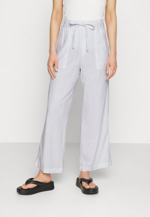 Vrouw draagt losse lichtgrijze broek met trekkoord en zijzakken, gecombineerd met zwarte platformsandalen, staand tegen een effen witte achtergrond.