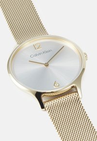 Montre avec bracelet en maille dorée et cadran rond argenté, dotée d'index dorés aux positions 12 et 6, et du logo "Calvin Klein" au centre.