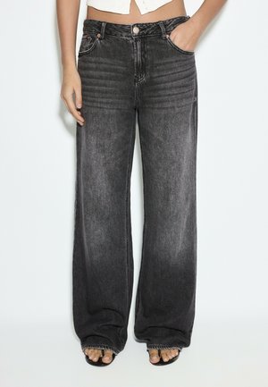 Person trägt hoch taillierte, weit geschnittene schwarze ausgewaschene Jeans mit weißem cropped Top, eine Hand in der Tasche und schwarze Sandalen, die Zehen zeigen.