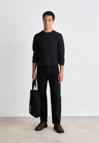 Camisa negra de manga larga con un pequeño logo, combinada con pantalones negros y zapatos de cordones marrones. Sosteniendo una bolsa tote negra con dos asas.
