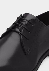Chaussure habillée en cuir noir poli avec laçage fermé et fines lacets ronds noirs sur fond blanc uni.