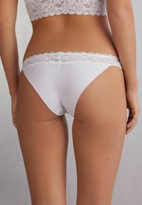 Culottes blanches avec des bordures en dentelle, un corps en tissu lisse et une coupe haute. La dentelle présente un motif géométrique le long de la taille.
