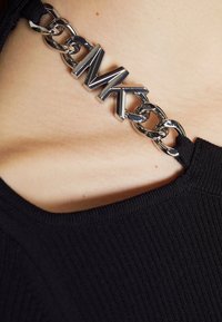 Haut noir côtelé avec une chaîne en argent présentant un logo "MK" au niveau de l'encolure. La texture du tissu semble douce.