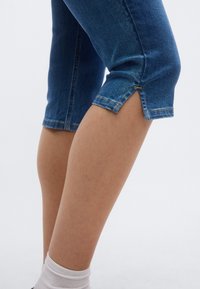 Τζιν καπρί σε σκούρο μπλε, με κοντό τελείωμα και χρυσές ραφές ως διακοσμητικά στοιχεία. Διαθέτει σχισμή στο τελείωμα για επιπλέον σχεδιαστική λεπτομέρεια.