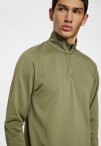 Olivgrön kvart zip-pullover med texturerade paneler, med hög krage och kontrasterande sömmar. Lätt material, mjuk struktur.
