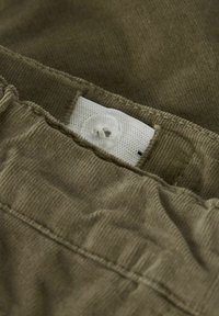 Tissu en velours côtelé vert olive avec une surface texturée, comportant une étiquette blanche cousue à l'intérieur et une fermeture à bouton translucide.