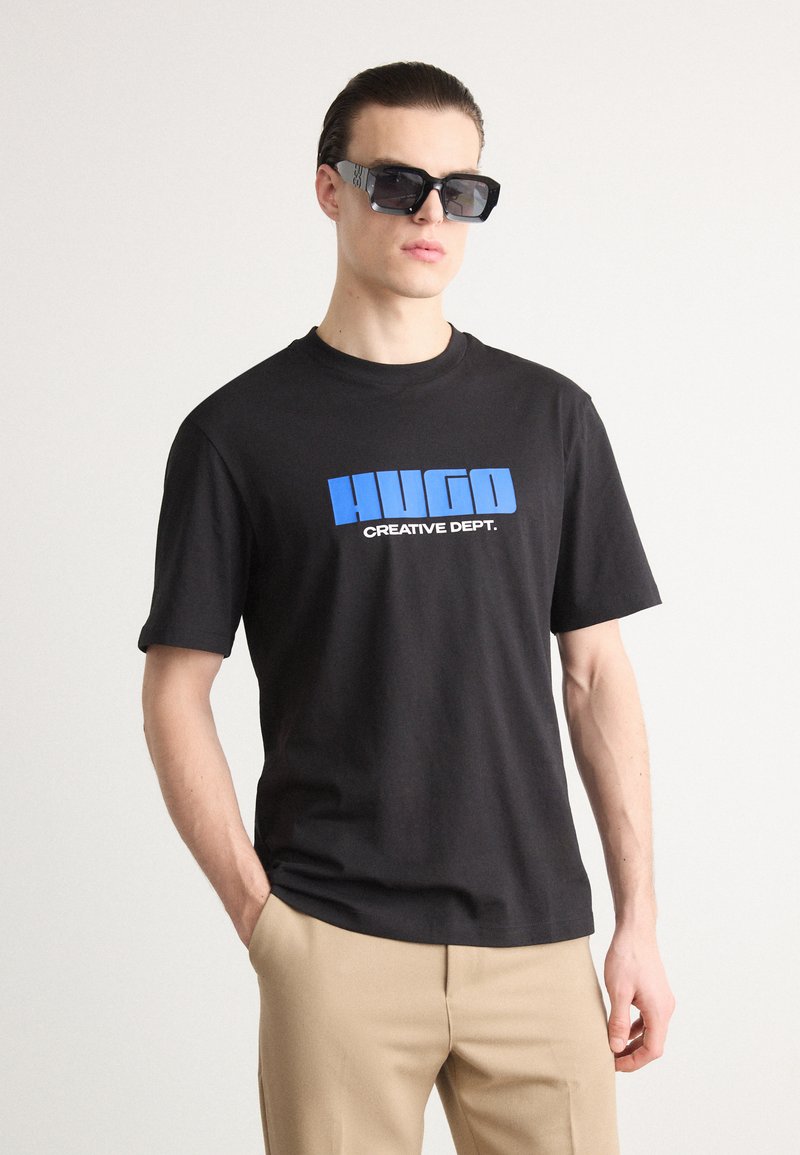 Svart bomull T-skjorte med en blå "HUGO" grafikk og teksten "CREATIVE DEPT." Modellen har på seg oversized svarte solbriller og beige bukser.