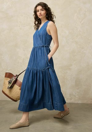 Frau mit lockigem Haar, die ein ärmelloses blaues Jeans-Wickelkleid und gewebte flache Schuhe trägt, hält eine große beige-braune Tasche mit einem Schal.