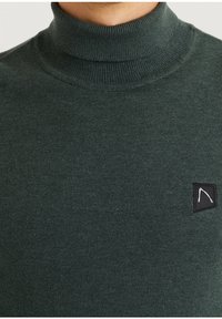 Dunkelgrüner Rollkragenpullover mit einer gerippten Struktur und einem kleinen schwarzen Etikett mit einem weißen Muster auf der Brust.