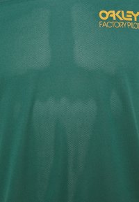Zelené tričko z síťoviny s nápisem „Oakley Factory Pilot“ ve žluté barvě. Má prodyšnou strukturu a jednoduchý design.