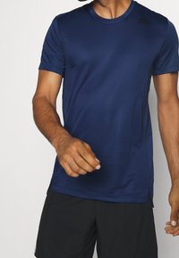 Muž oblečený v námořnicky modrém sportovním tričku Adidas a černých šortkách, stojící na jednobarevném světlém pozadí.