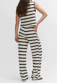 Femme debout avec une main sur la hanche, portant un haut en tricot sans manches noir et blanc à rayures horizontales et un pantalon large assorti avec des talons.