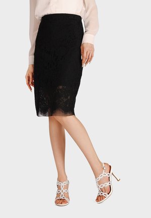 Donna con una gonna nera in pizzo fino al ginocchio, una camicetta trasparente e sandali bianchi con tacco alto e cinturini decorativi, in piedi con una gamba incrociata.