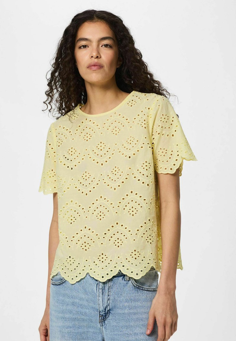 Femme aux cheveux bouclés portant un chemisier en dentelle ajourée jaune pâle avec des bords festonnés et un jean en denim bleu clair, debout sur un fond blanc.