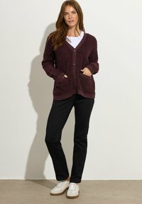 Bordeaux, puszysty sweter z dekoltem w serek, z przodu na guziki, dwie boczne kieszenie i ściągacze przy mankietach, w połączeniu z czarnymi spodniami i białymi trampkami.