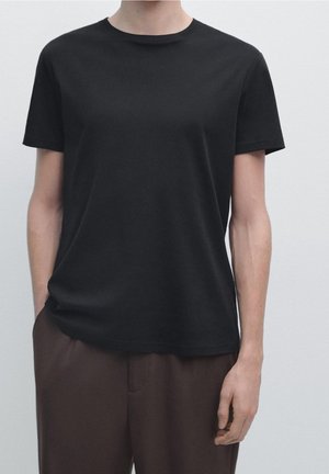 Persona che indossa una semplice t-shirt nera a maniche corte e pantaloni marroni in piedi davanti a uno sfondo grigio chiaro.