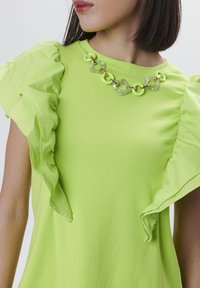 Top verde lime con maniche arricciate, realizzato in tessuto liscio. Presenta una collana con catena spessa con anelli trasparenti e verde lime.