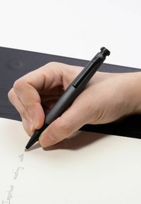 Mustekynä, jota pidetään kädessä, kirjoittaa vaaleankeltaiselle paperille. Taustalla on tumma, sileä pinta.