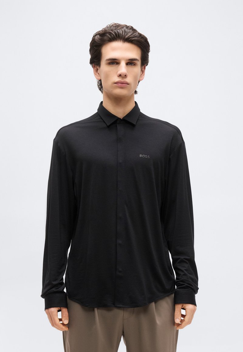 Νεαρός άνδρας με σκούρα μαλλιά, που φοράει μαύρο μακρυμάνικο πουκάμισο με κουμπιά και μικρό λογότυπο "BOSS" και μπεζ παντελόνι, στέκεται μπροστά σε απλό φόντο.