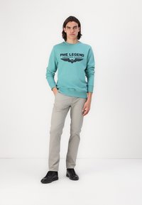 Svjetloplava dukserica s crnim grafikonom "PME LEGEND", u kombinaciji sa svijetlosivim hlačama i crnim tenisicama. Jednostavan dizajn, ležeran kroj.