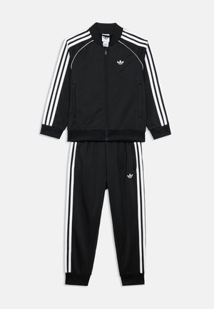 Mustast siledast kangast dressipluus, millel on valgete triipudega tõmbelukuga jakk ja sobivad püksid elastse vöökoha ning taskutega.