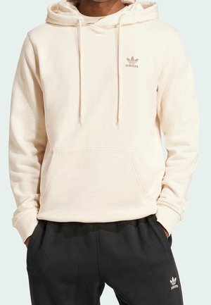 Mężczyzna w kremowej bluzie z kapturem Adidas z kieszenią z przodu i czarnych spodniach dresowych Adidas, ręce w kieszeniach, na jednolitym tle.