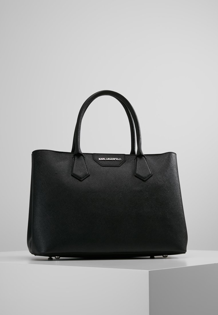 Bolso tote negro con superficie texturizada, asas dobles y acentos angulares. Cuenta con una placa con el logo en la parte frontal. Construcción robusta con un diseño espacioso.