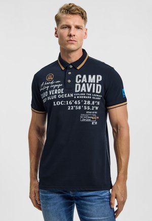 Man wearing navy blue polo shirt with sailing-themed text and coordinates, paired with blue jeans, standing against a plain light background.