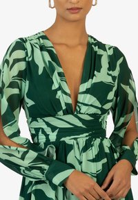 Robe florale verte avec un décolleté en V profond, des manches longues en tissu transparent et une taille cintrée. Tissu texturé avec une jupe fluide et des détails accentués.